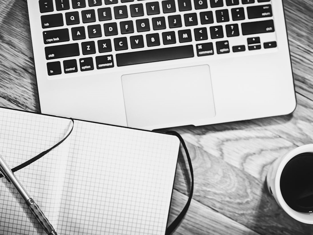 Grayscale picture of laptop, notebook, pen and cup of coffee lying on a table